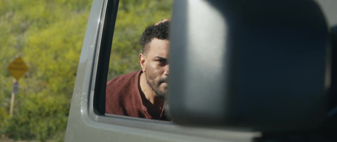 A man leans over and fixes his hair looking in a large truck side mirror