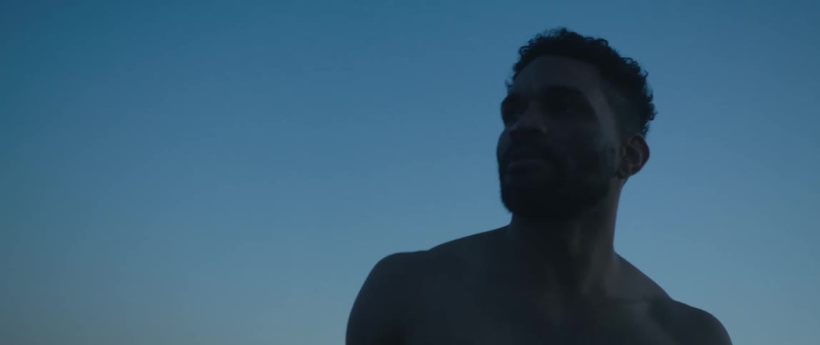 Shirtless man with beard is silhouetted against the sky at dusk