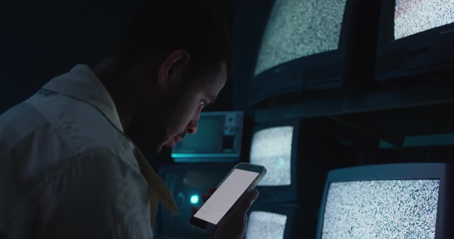 A man is holding a phone and looking at static on the screen and there are several monitors on the wall near him with static on all the screens