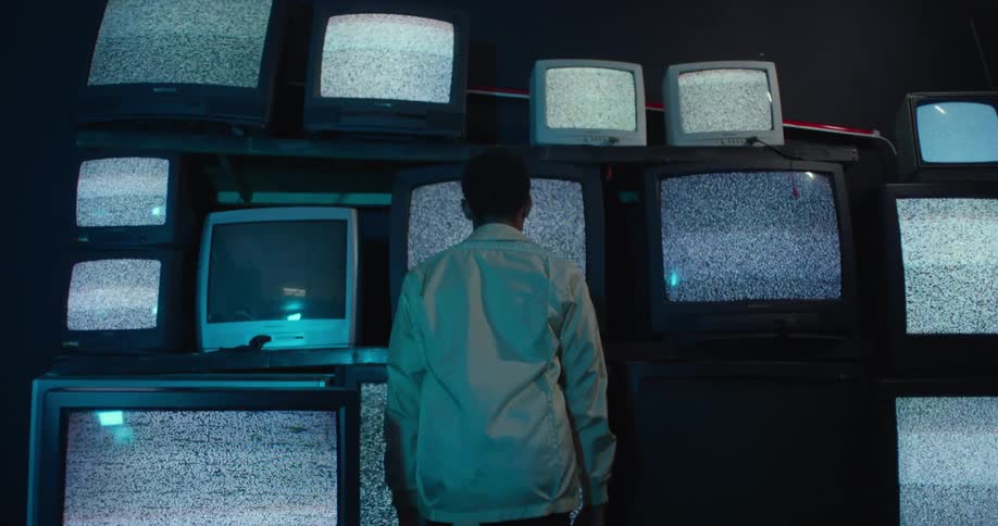 A man is sitting in front of a wall of televisions with static on the screens