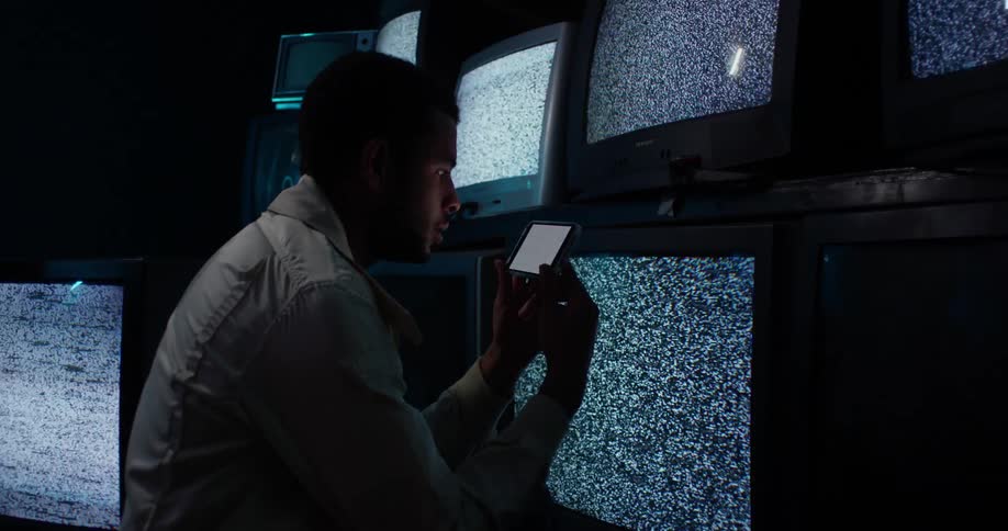 A man is holding a phone and looking at static on the screen and there are several monitors on the wall near him with static on all the screens