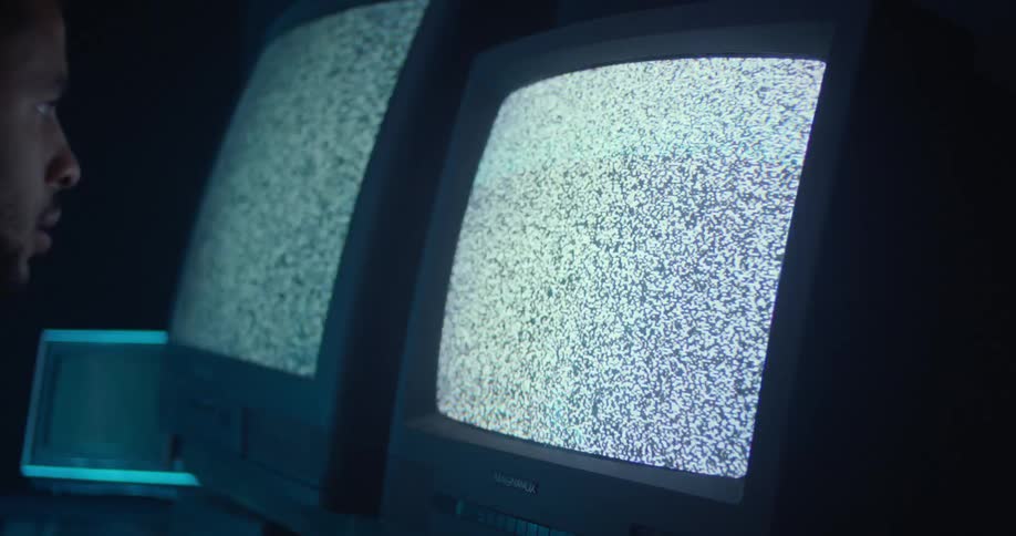 A man stares into the screen of an old television displaying static as he slowly backs away in a dark room