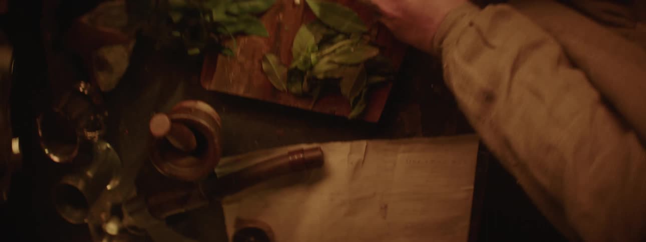 Man slices herbs with a knife over a board inside a medieval kitchen