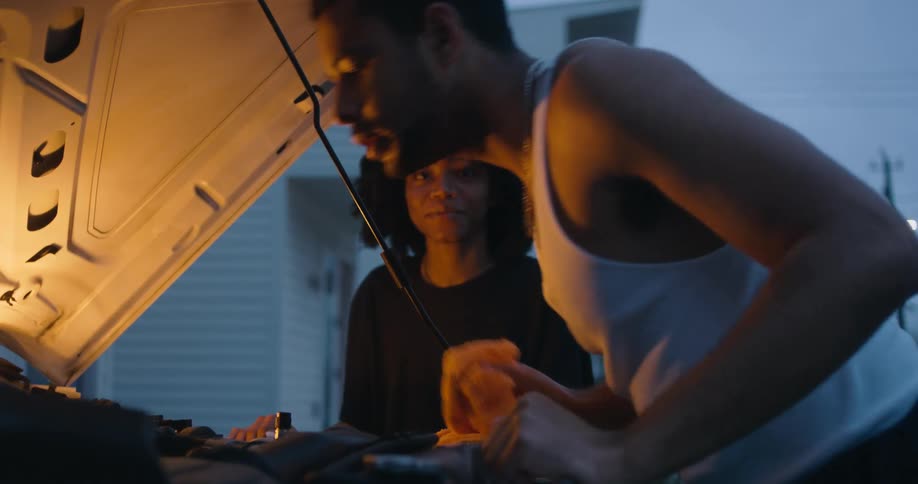 A woman talks to a man as he examines the engine of a car in the evening