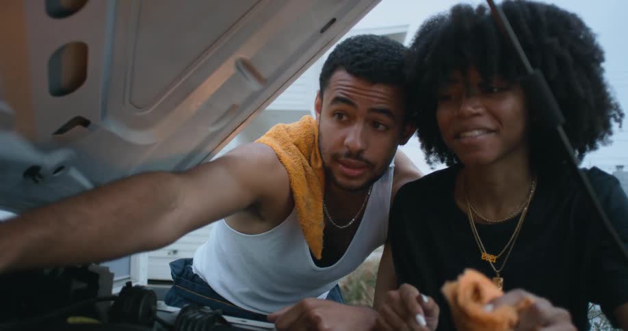A man and woman talk with one another while examining the engine of a car parked in a driveway outdoors during the day