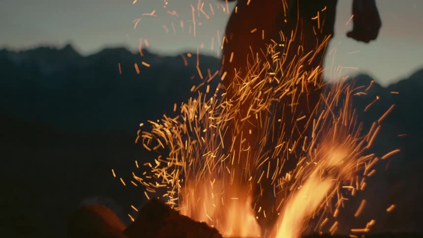 A person is standing outdoors by a bonfire at dusk