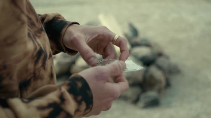 A man struggles to use a compass while standing next to an unlit campfire in the desert