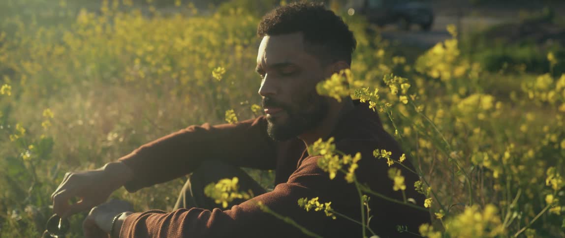 Young man sitting on a field of yellow wild flowers takes sunglasses out in the morning