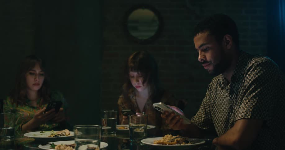 A group of people sitting together at a dining table all look at their smartphones while ignoring one another