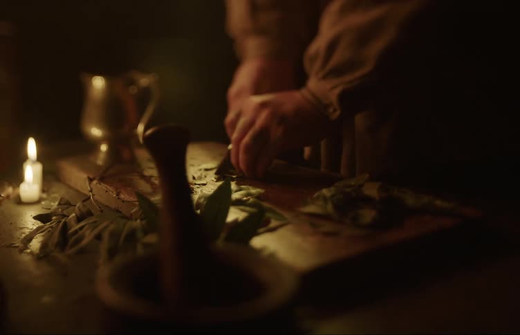 Man slices herbs with a small knife over a wooden table in candlelight at night