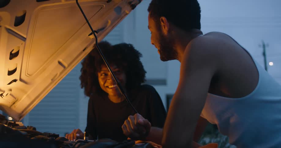 A woman and man talk with each other while examining the engine of a vehicle outdoors in the evening