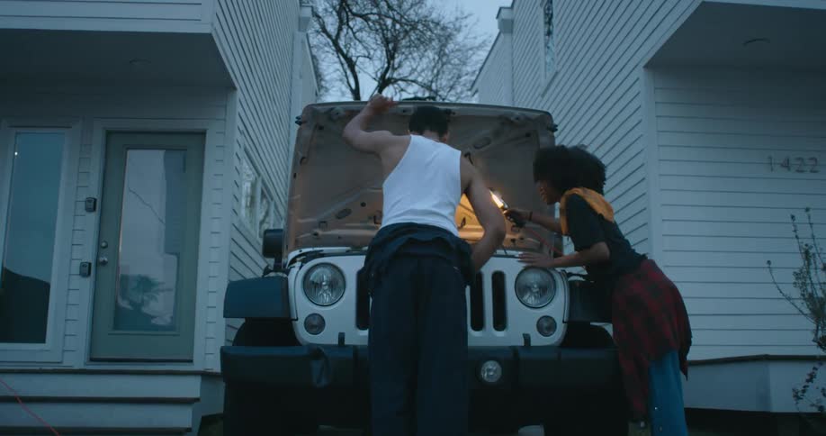 Young couple checking the engine of a jeep parked between houses at evening