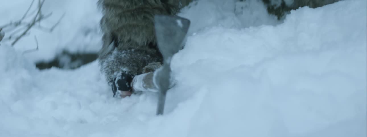 A hand slams a stake into the snow and the camera pans up to reveal a bearded man in heavy furs who is breathing hard and struggling to pull himself forward