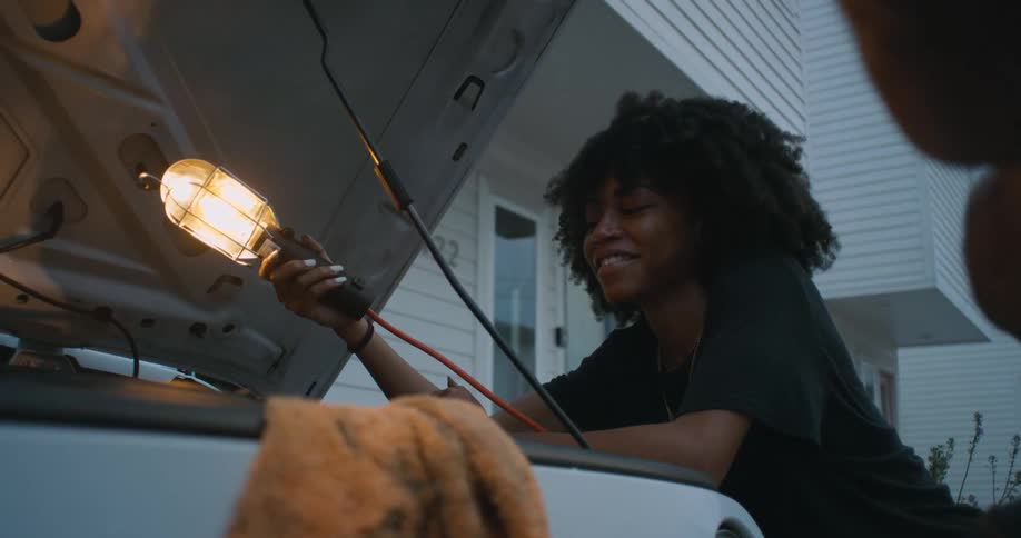 Woman holds car work light over open engine compartment of vehicle parked near house