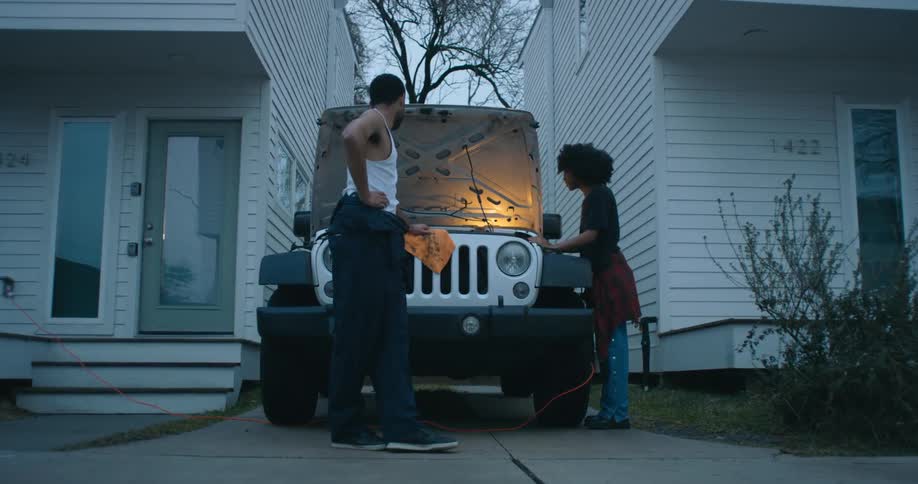 A man and woman both working under the hood of a jeep in a driveway