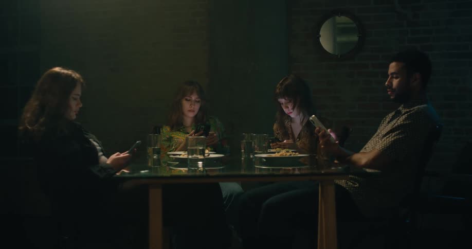 A group of friends are eating dinner at the table and are on there phone