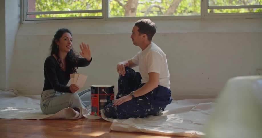 Man and woman sit on drop cloths in wood floored room near a large can of paint and discuss paint samples
