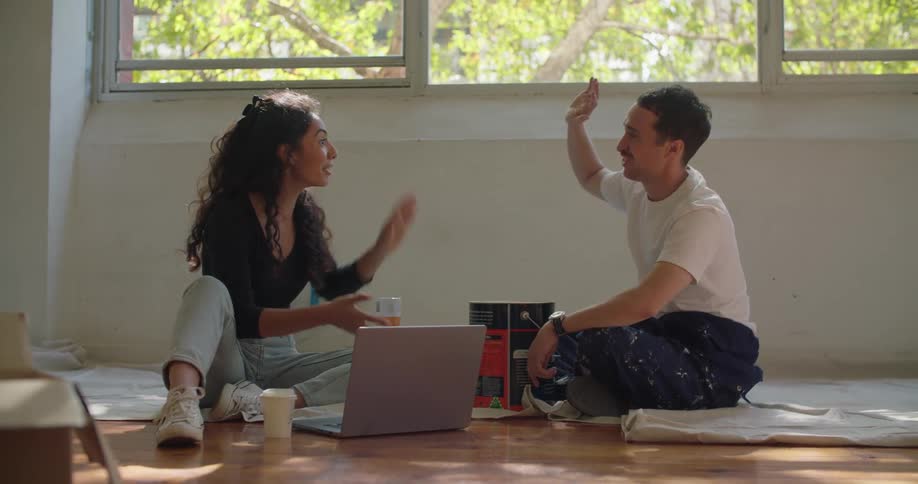 Man and woman sit on drop cloths near can of paint and laptop talking before sharing a high five