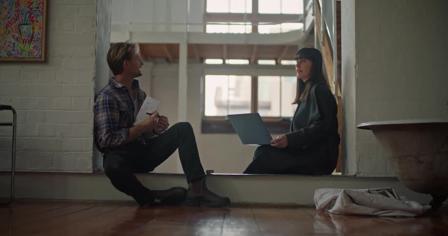 A woman holding a laptop sits on the floor of a house and talks to a man as he takes notes