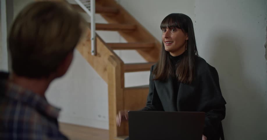 Female real estate agent with forelock hair and black overcoat talks with male customer in the empty building