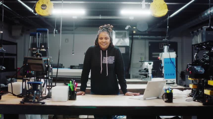 A woman smiles into the camera while standing behind a table in a workshop