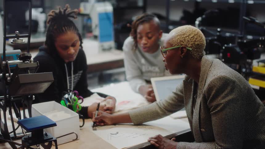 Two teenage girls lean over a desk and listen to a professor as she sketches and talks with them