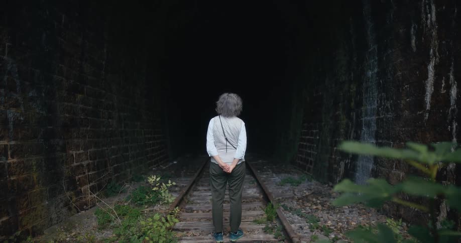 An elderly woman stands on train tracks and looks down a tunnel
