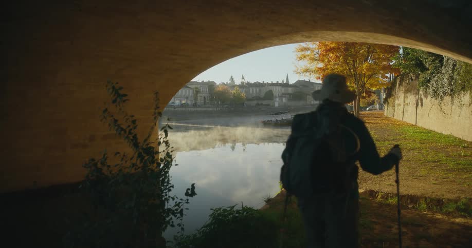 A hiker hikes beneath a bridge over a river running through a town