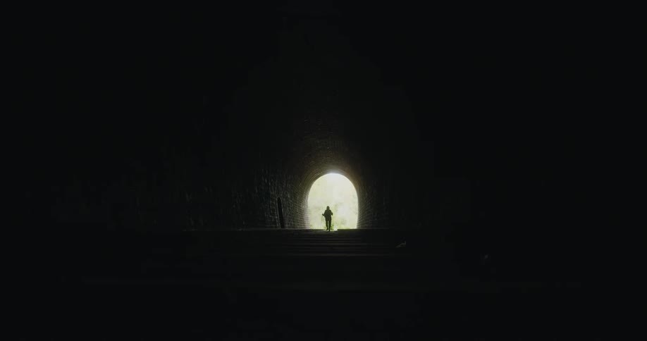 A hiker walks along a railway through a dark tunnel towards the opening on the other side