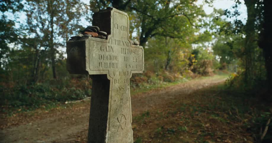 A cross shaped gravestone with flat rocks sitting upon it near a dirt road and trees are in the distance