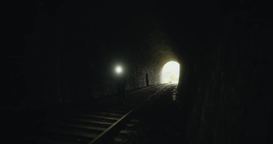 A person shines a flashlight while walking down train tracks in a dark tunnel