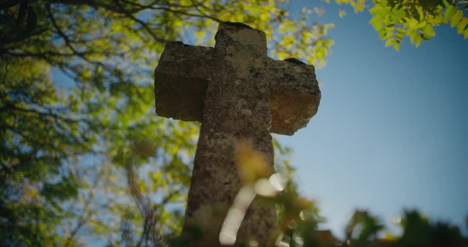 The sun shines on a cross shaped gravestone and trees are in the distance
