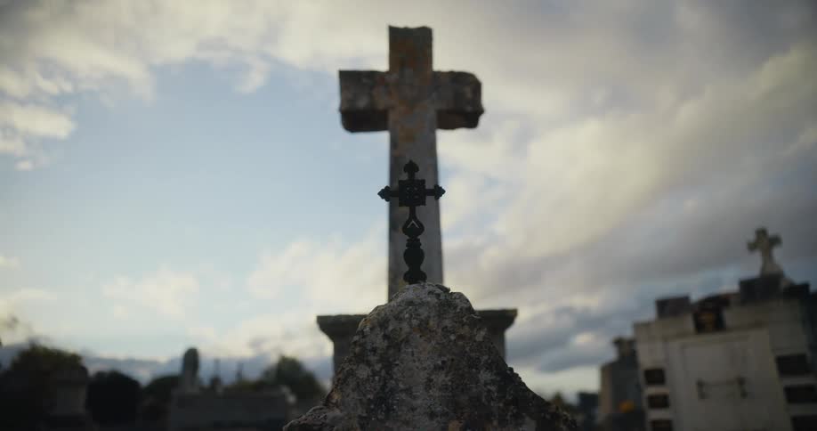 Worn headstone and stone cross sit in graveyard near crypt on cloudy day