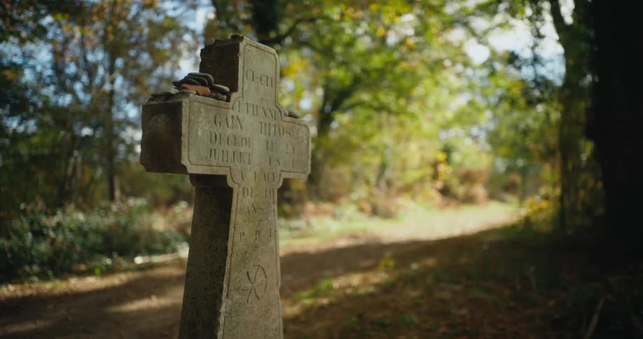 The sun shines on a cross shaped gravestone with flat rocks sitting upon it and trees are in the distance