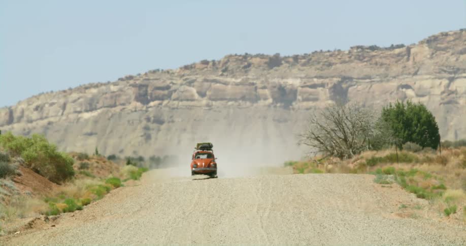 Red vw bug drives along dirt road passing scrub with cliff in the background