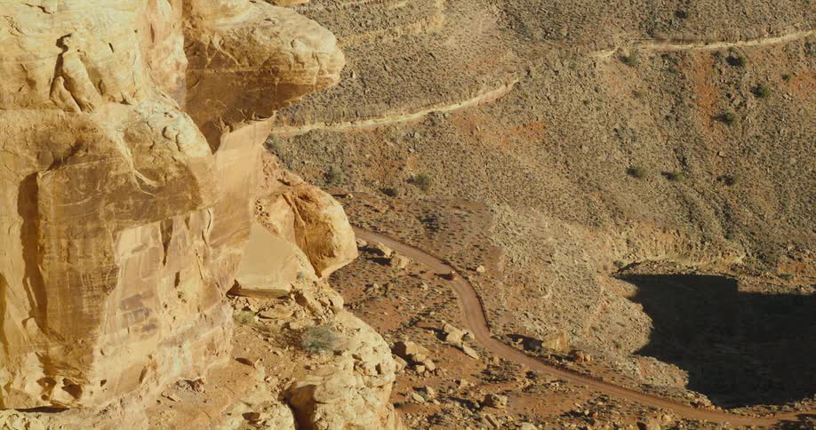 A vehicle travels along a dirt road beneath a cliff in an arid countryside during the day