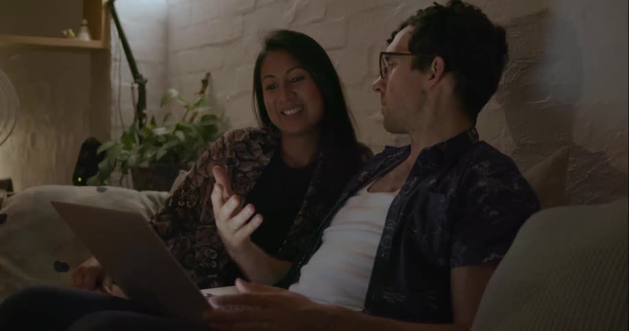 A man and woman sit on a couch talking and looking at a laptop