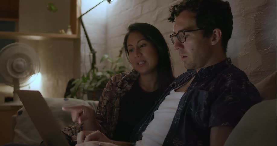 Man and woman sit on a couch talking and looking at a laptop