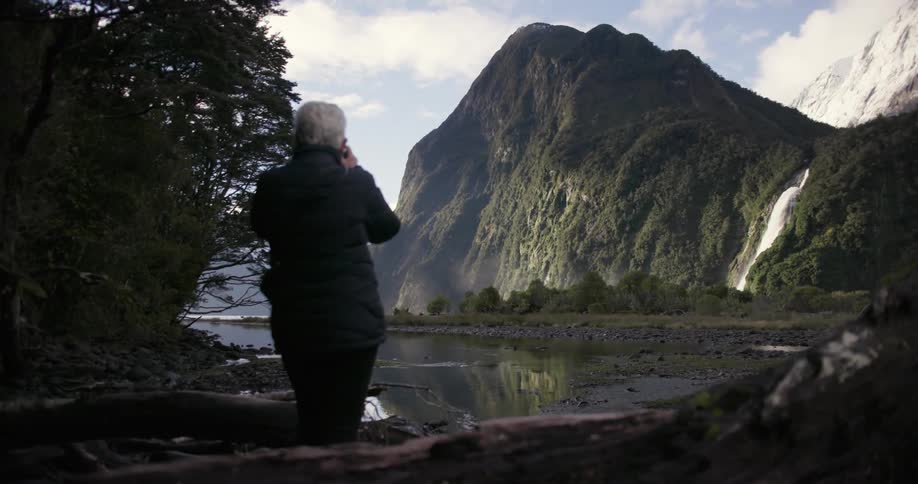 A person takes a photograph of a lake near a mountain