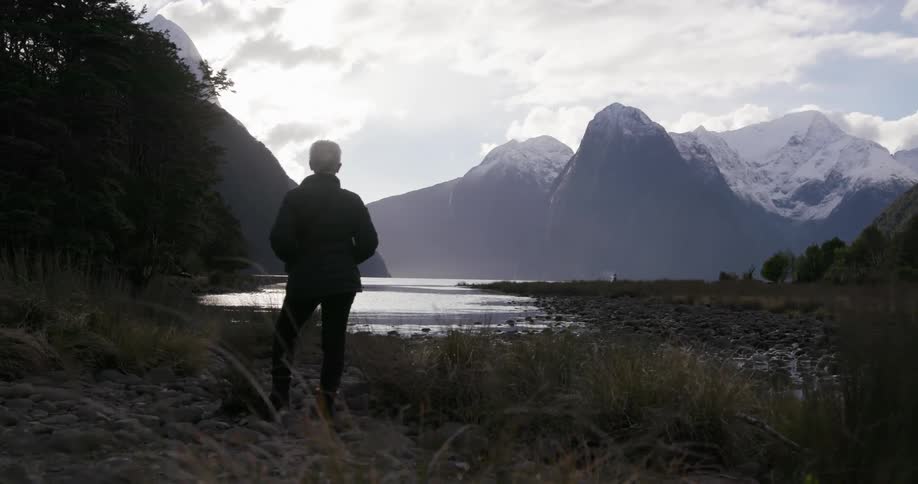 A woman stands in front of a flowing lake in front of mountains
