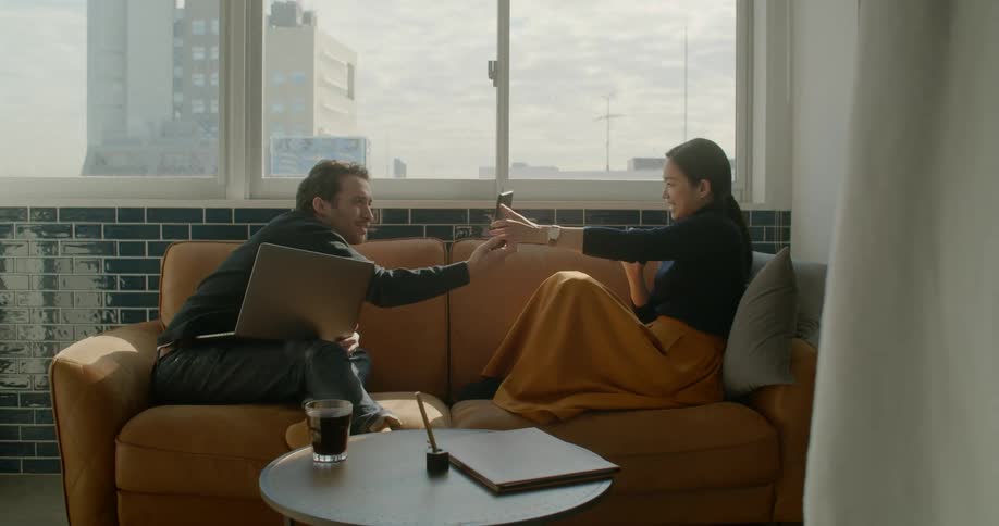 A woman shows her cellphone to a man as he uses his laptop