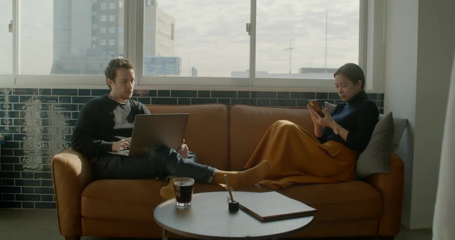 White man and asian woman sitting on the sofa uses electronic devices