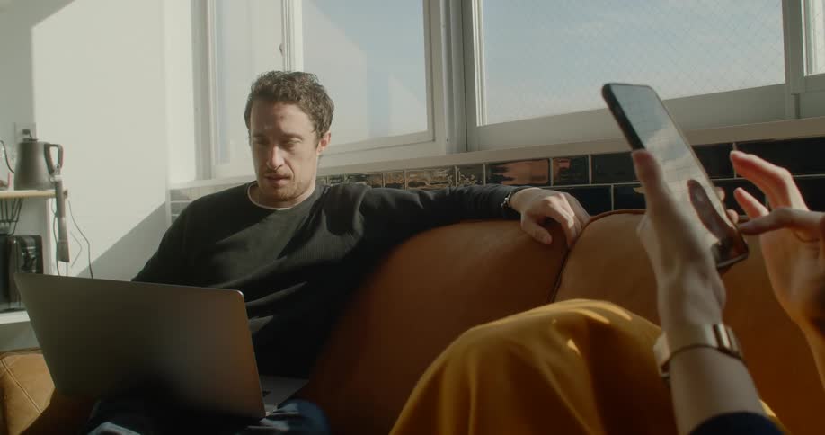 A man sits on a couch with a laptop beside a person on their cell phone