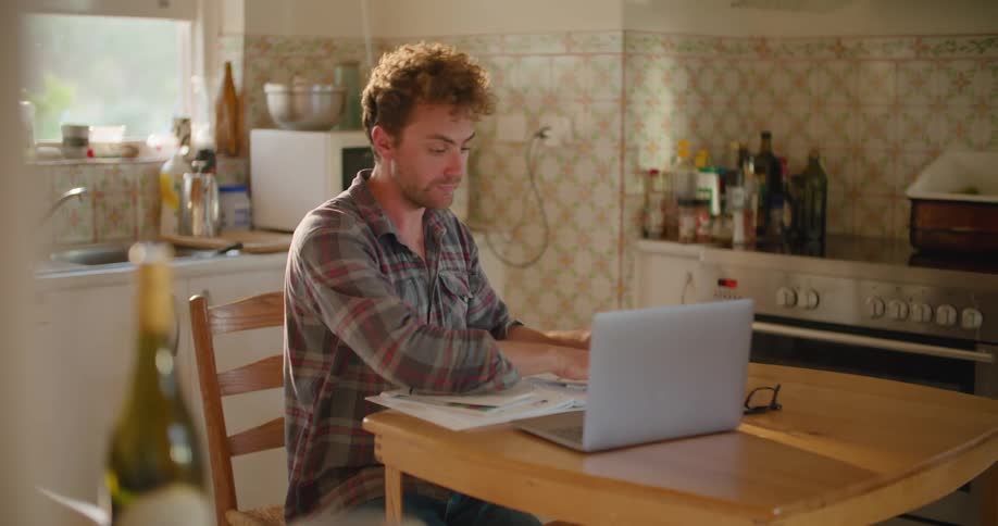 A man sitting at a table in a kitchen and looking at a laptop with a wall and window in the background