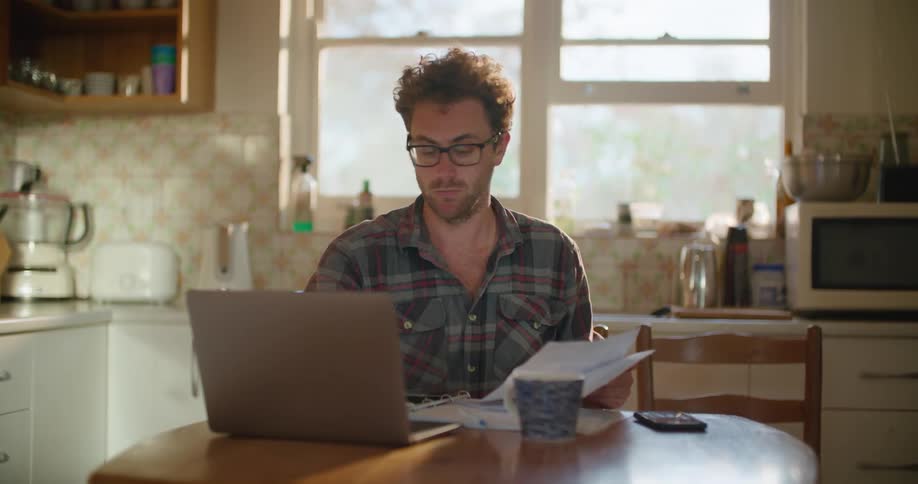 Man looks at some papers while working on a laptop in the kitchen