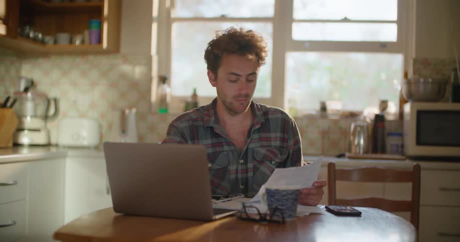 A man is sitting at a kitchen table reading paperwork and reading on a laptop