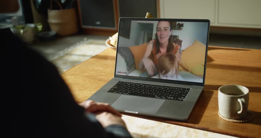 A woman holds a child visible on a laptop screen communicating with a man sitting on a couch in front of the laptop