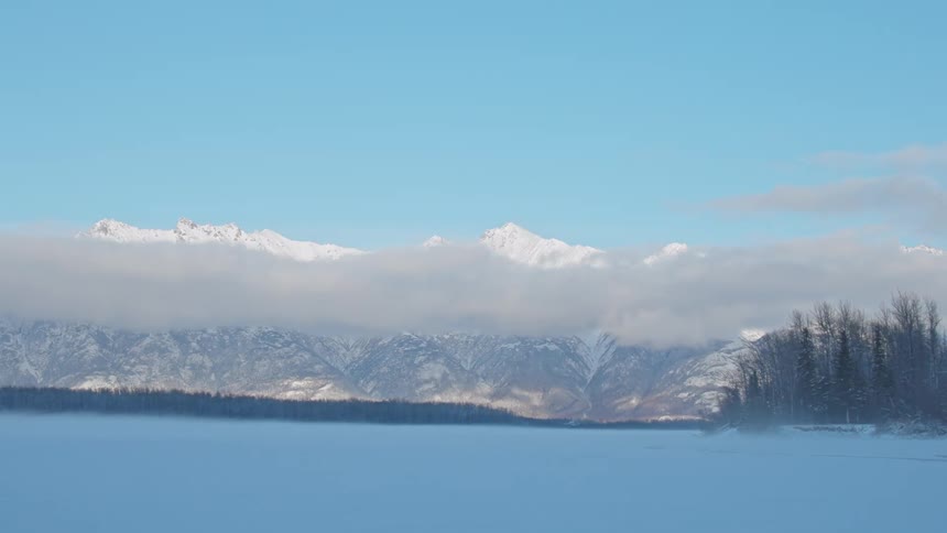 Snow blows across snow covered area stretching to conifer forest and mountains on the horizon