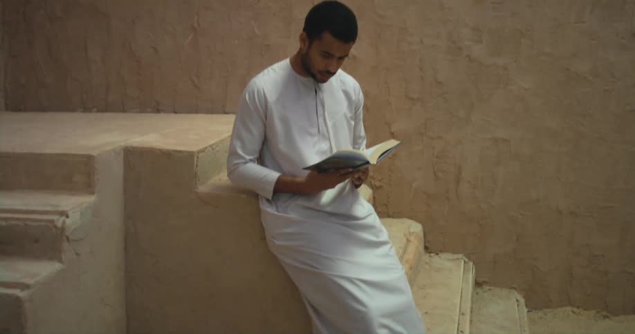 A man wearing a white tunic is leaning against stairs reading a book