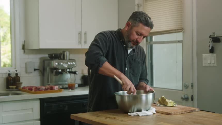A man stirs food in a mixing bowl with a whisk while standing in a home kitchen during the day
